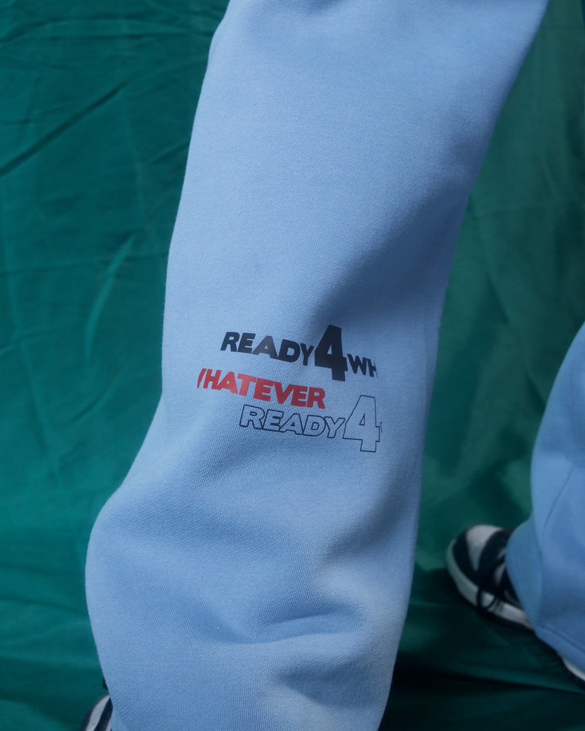 A close-up of a light blue sweatshirt or sweatpants leg with a printed graphic reading 'READY 4' and 'WHATEVER' in black and red text, set against a dark green background.