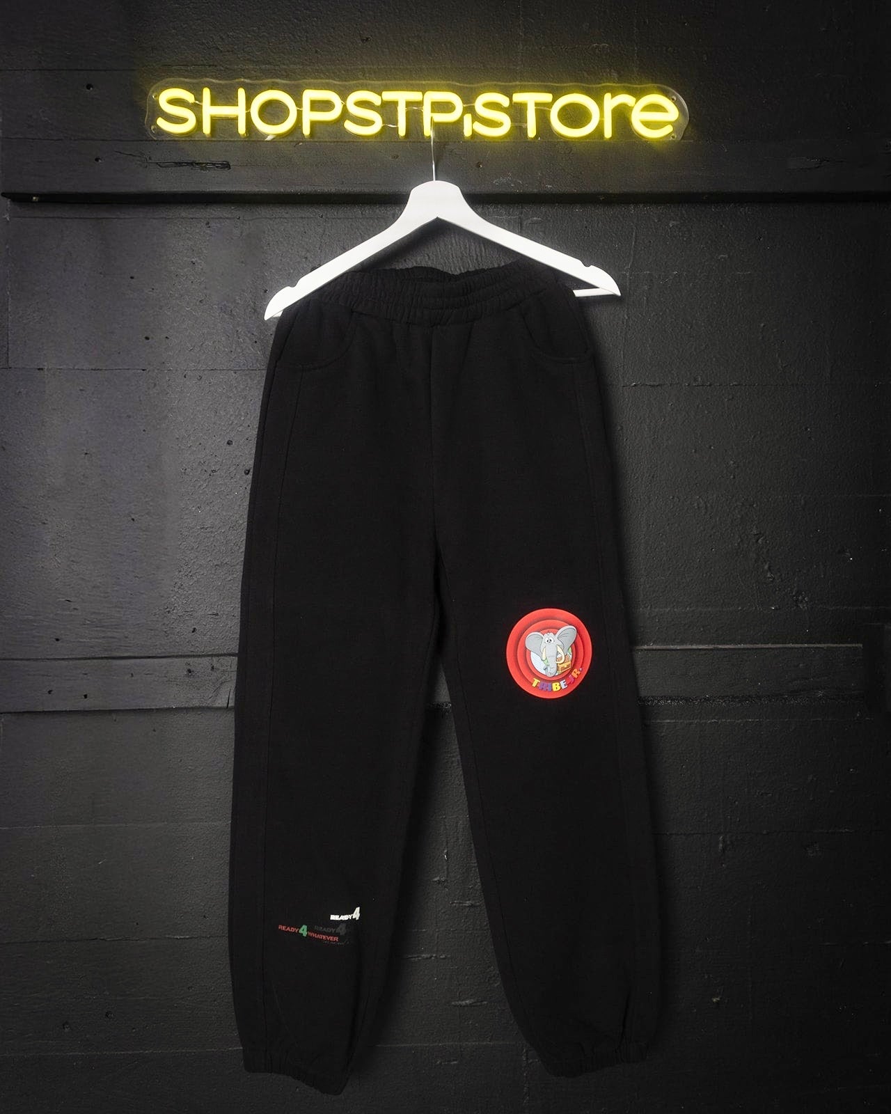 Black pants with a red patch on a hanger against a dark wall with 'SHOPSTPSTORE' neon sign.