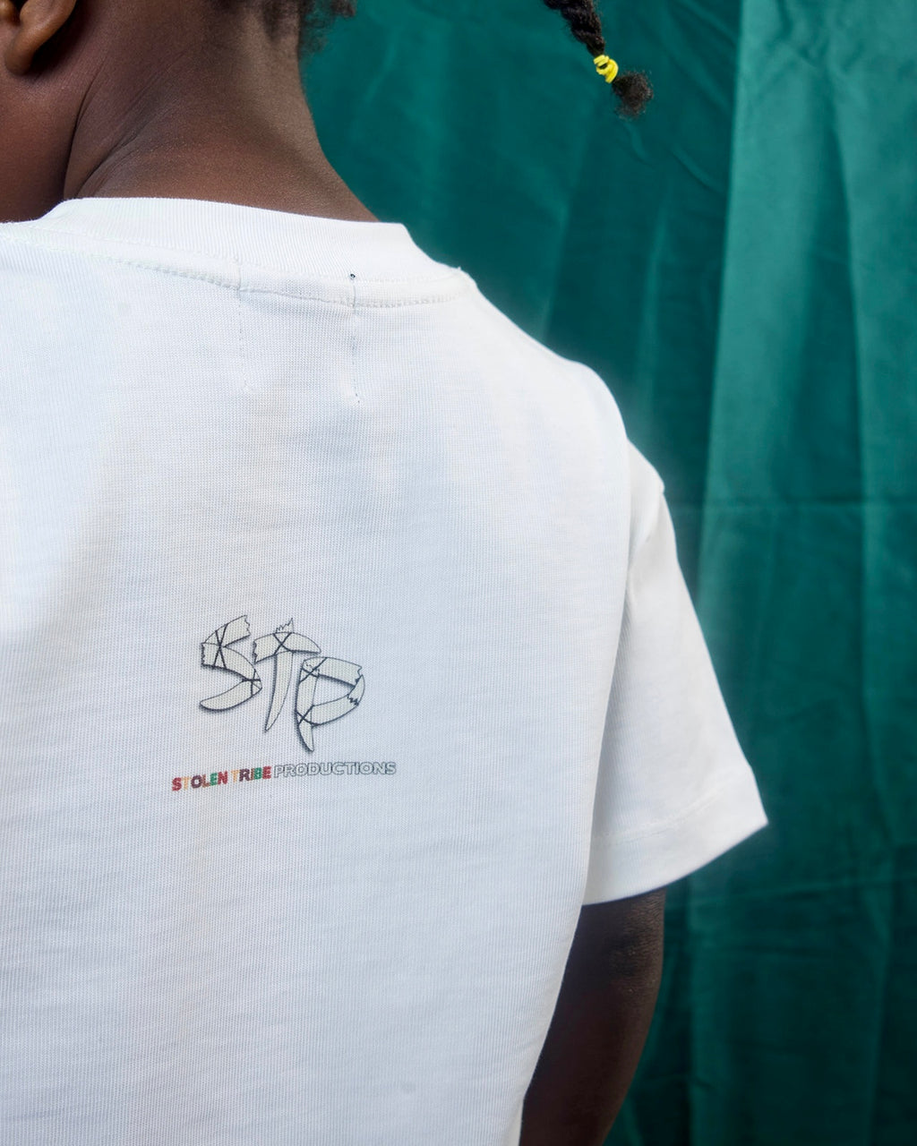 A close-up back view of a person wearing a white t-shirt with a black graphic and red text reading 'STP' Stolen Tribe Productions on the back, set against a dark green draped fabric background.