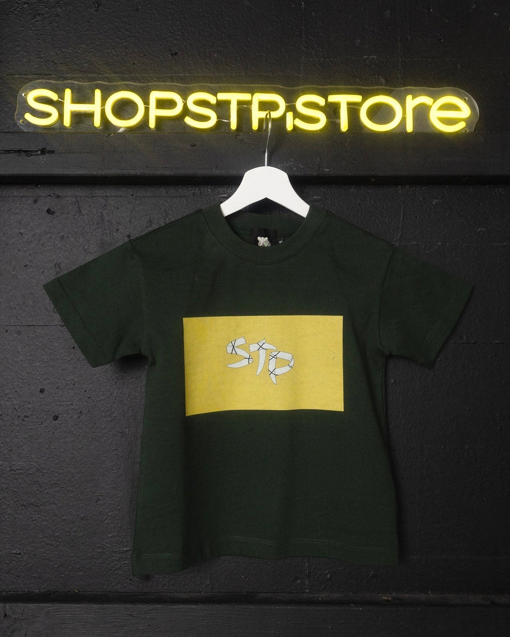 Green t-shirt with yellow logo on a hanger in front of a neon 'SHOPSTP:STORE' sign.