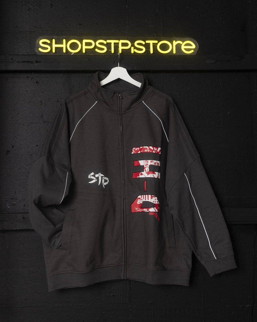 Gray jacket with red and white designs on a hanger against a dark background with 'SHOPSTPSTORE' neon sign.