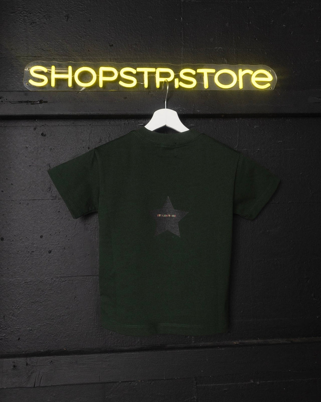 Green t-shirt with a star design on a hanger in front of a neon 'SHOPSTP.STORE' sign.