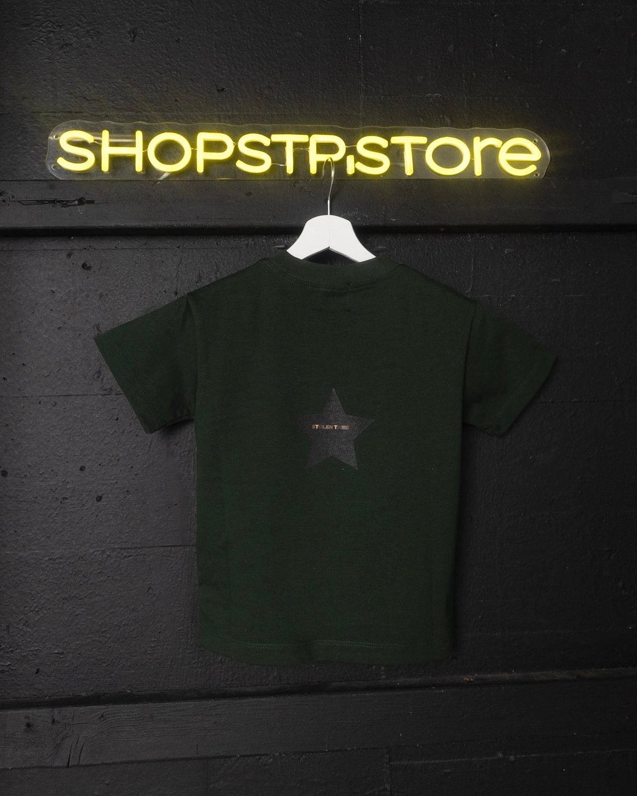 Green t-shirt with a star design on a hanger in front of a neon 'SHOPSTP.STORE' sign.