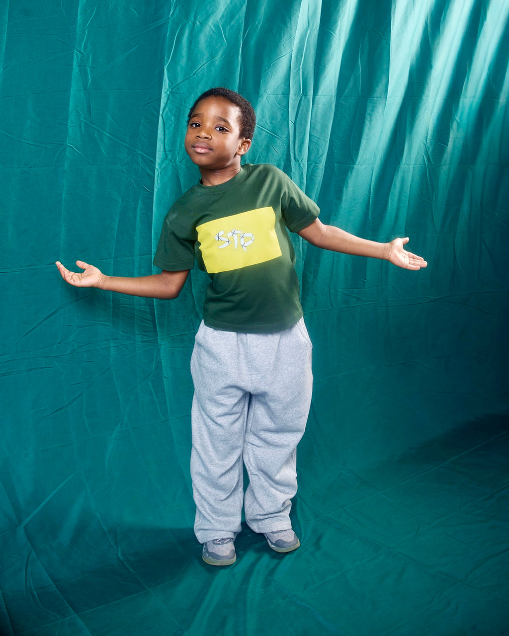 A young boy stands in front of a teal fabric backdrop wearing a dark green t-shirt with a yellow graphic that reads 'STP' and light gray sweatpants.
