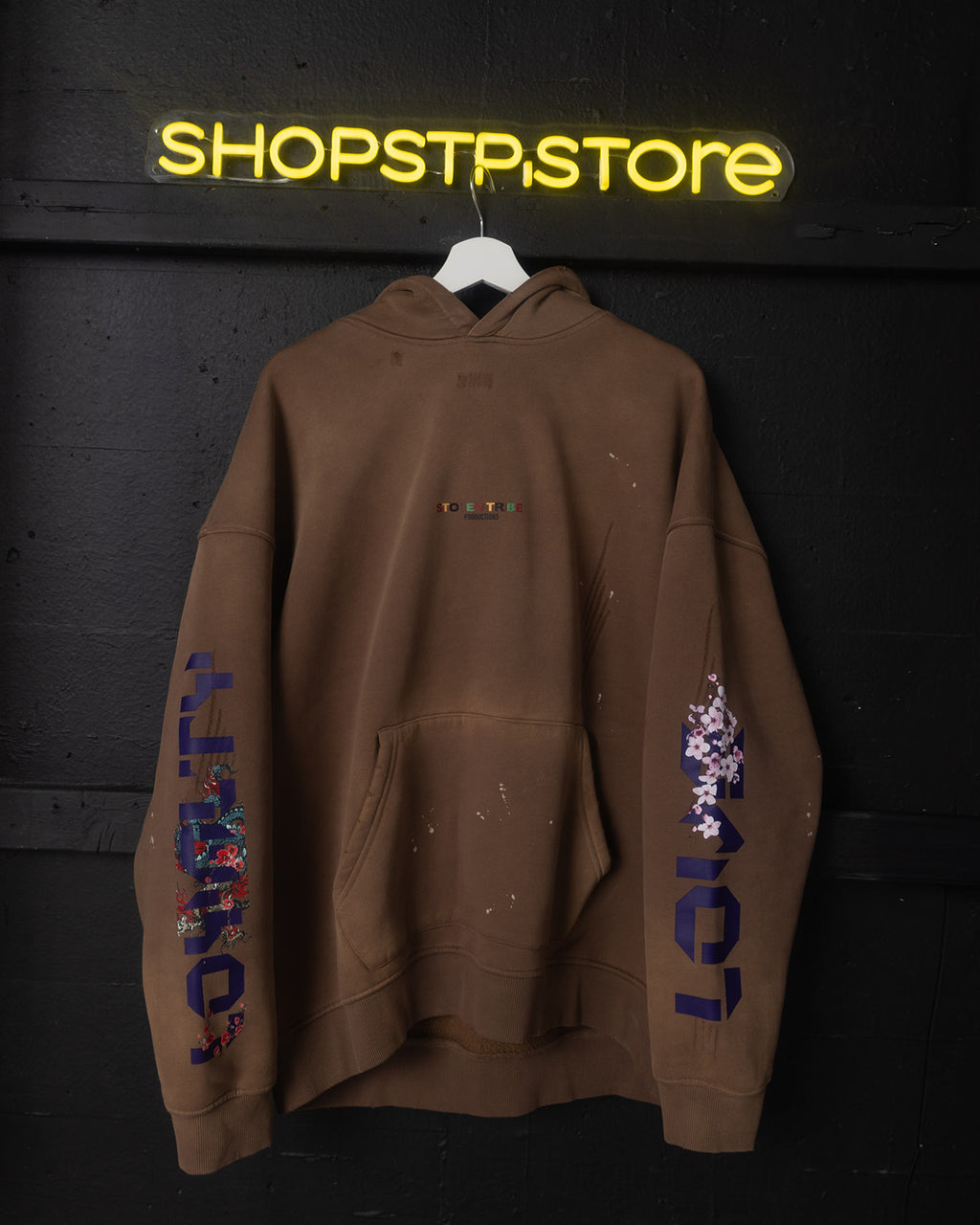Brown hoodie with colorful text and designs on a black background with 'SHOPSTP STORE' neon sign.