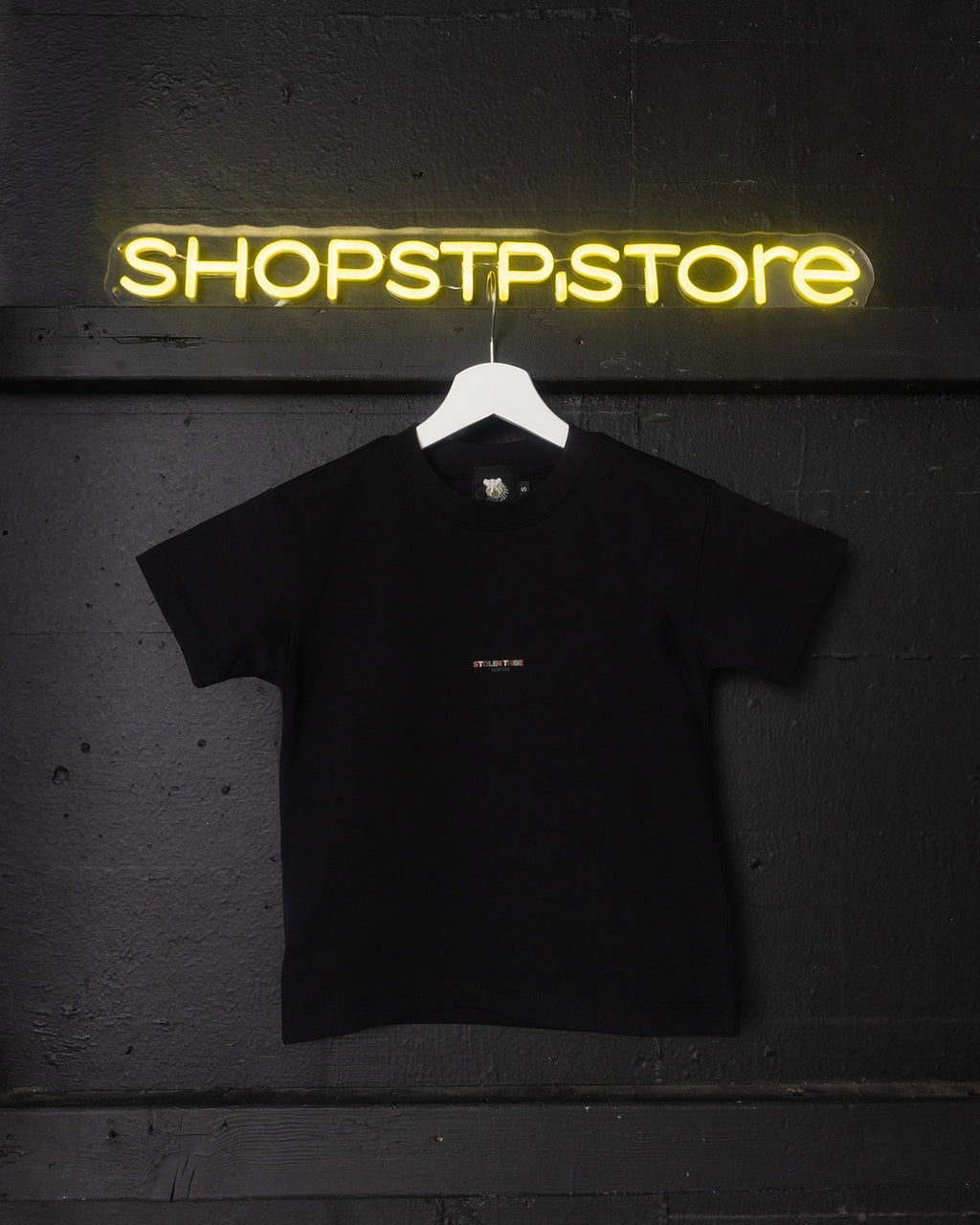 Black t-shirt with a small white logo on a hanger in front of a neon 'SHOPSTPSTORE' sign.