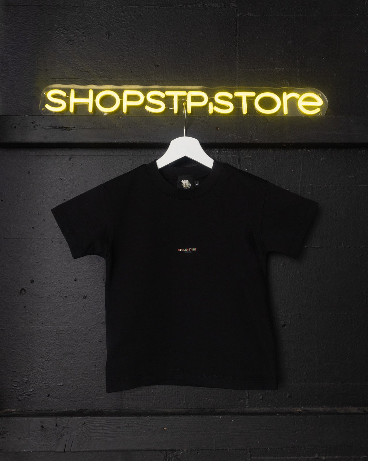 Black t-shirt with a small white logo on a hanger in front of a neon 'SHOPSTPSTORE' sign.