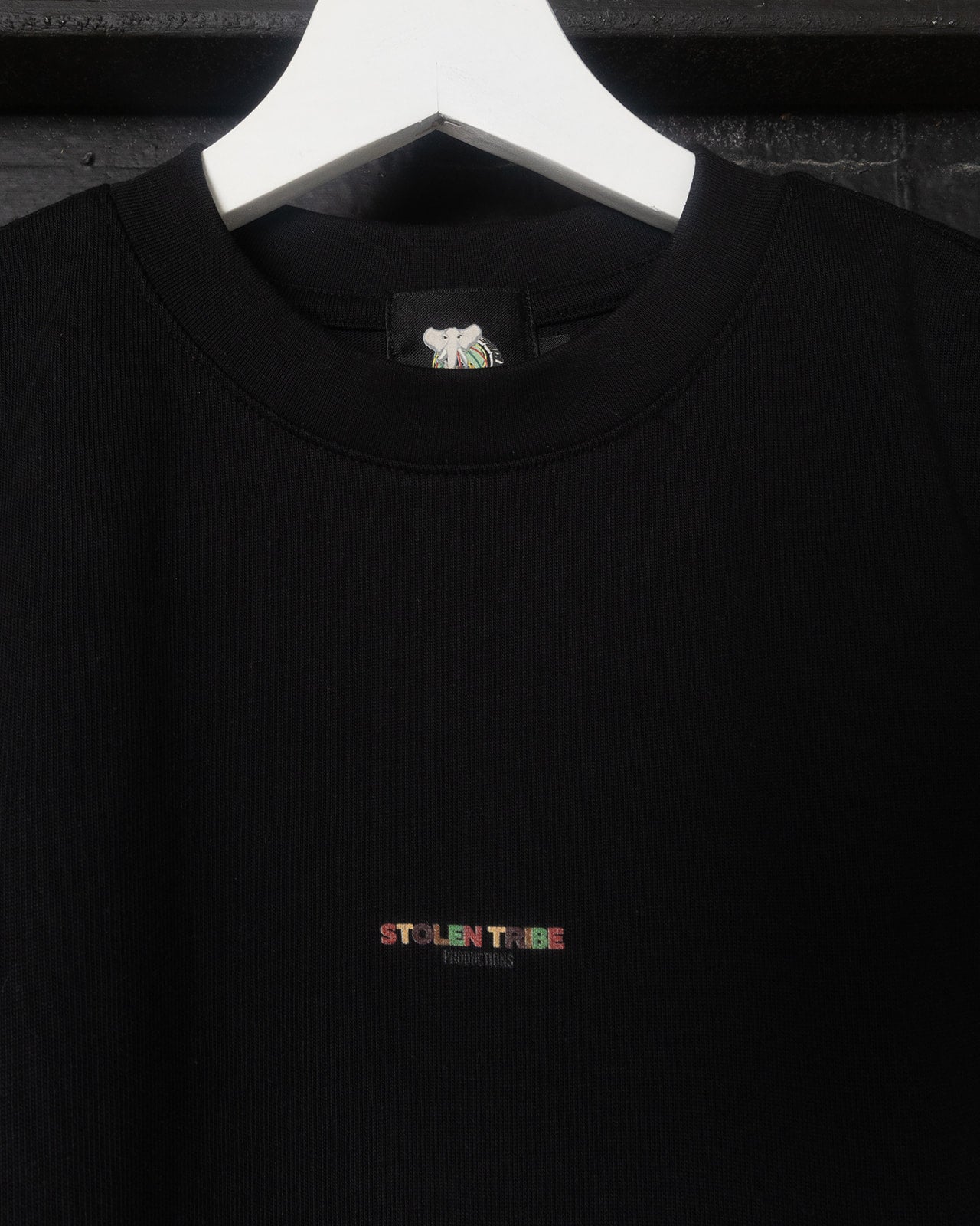 Black shirt with a colorful 'Stolen Tribe' logo on a white hanger against a dark background