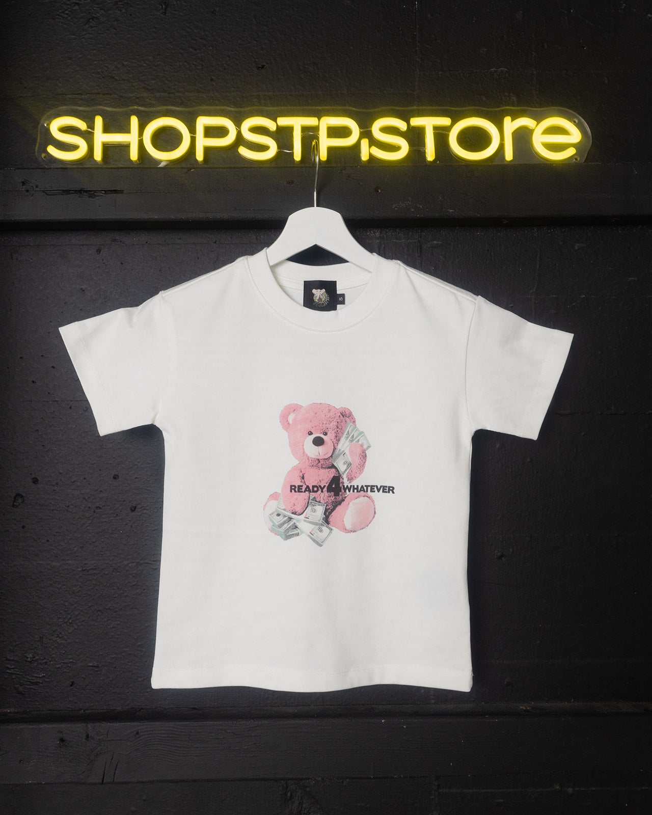 White t-shirt with a pink teddy bear graphic and text on a hanger against a dark background with 'SHOPSTP STORE' neon sign.