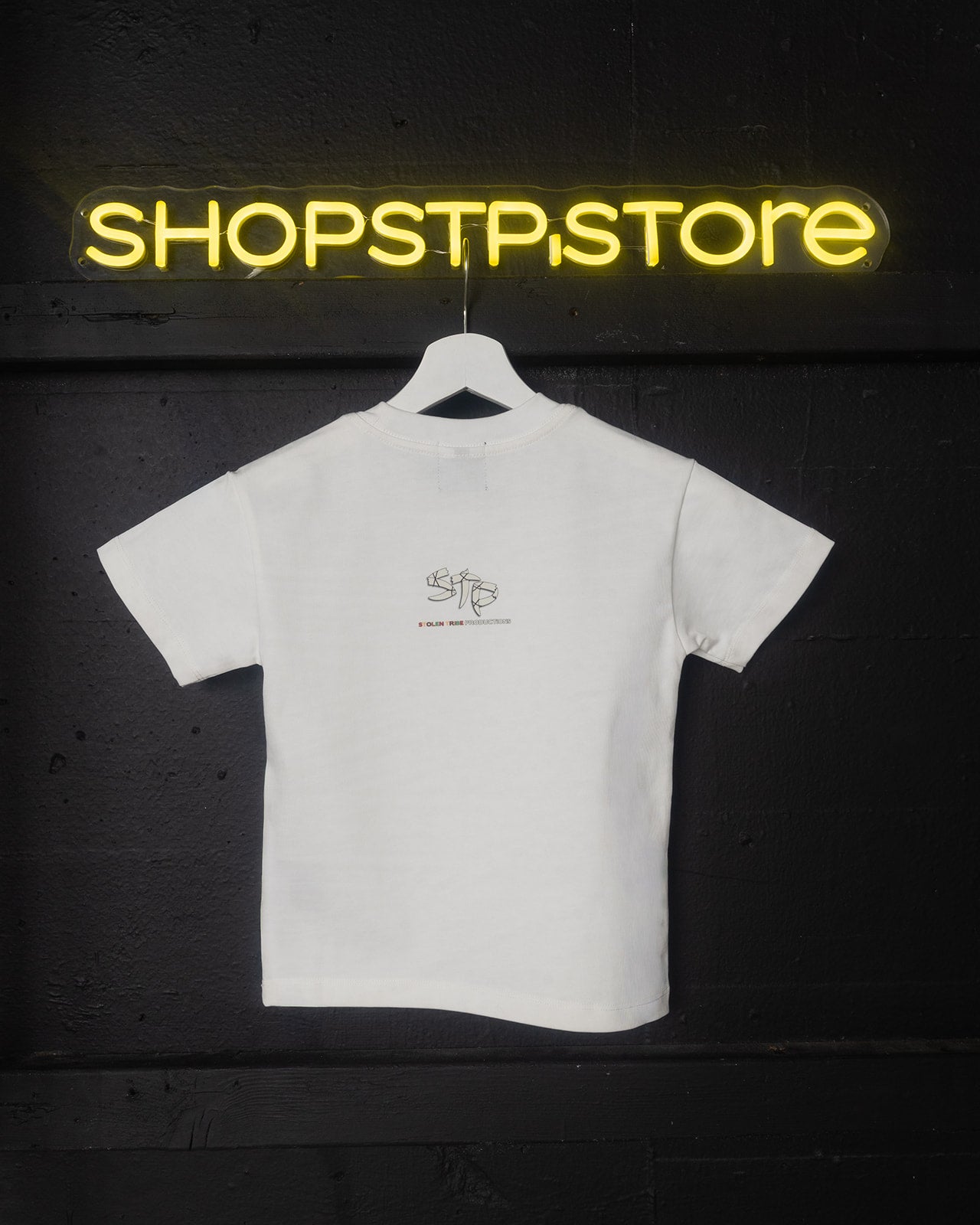 White t-shirt with a logo on a hanger in front of a neon sign reading 'SHOPSTPSTORE' on a dark background.
