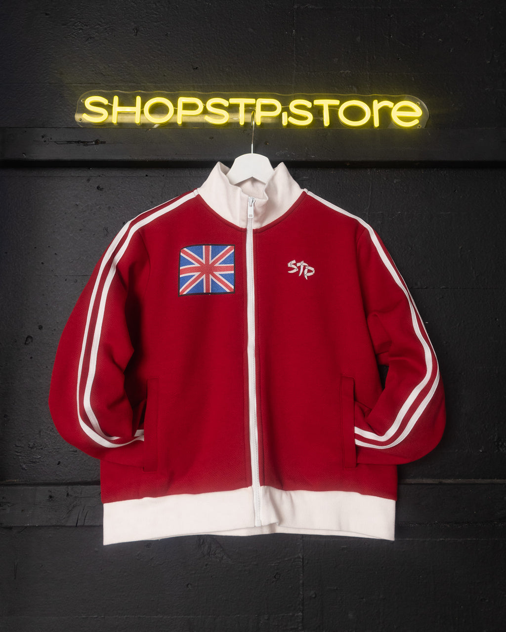 Red jacket with British flag patch and 'STP' logo on a hanger against a dark background with 'SHOPSTP.STORE' neon sign.