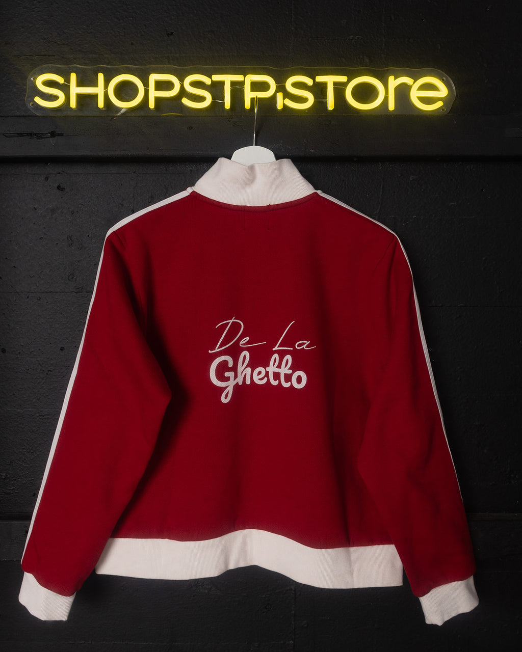 Red sweatshirt with 'De La Ghetto' text on a dark background with neon 'SHOPSTP STORE' sign.
