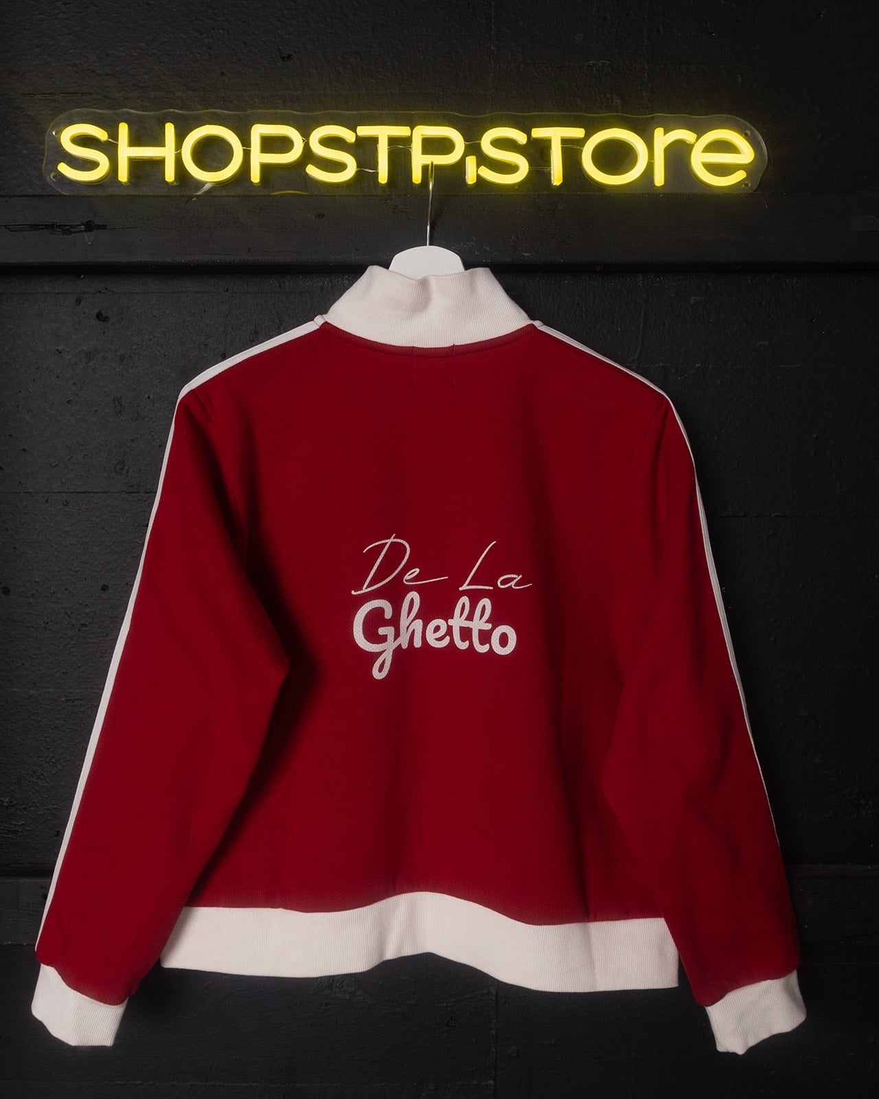 Red sweatshirt with 'De La Ghetto' text on a dark background with neon 'SHOPSTP STORE' sign.