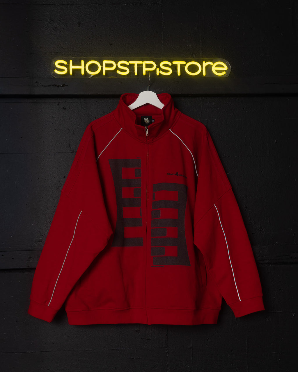 Red jacket with a logo on a hanger against a dark wall with 'SHOPSTP.STORE' neon sign.