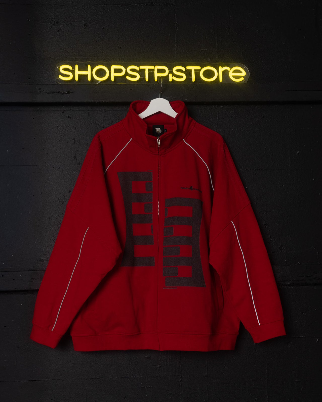 Red jacket with a logo on a hanger against a dark wall with 'SHOPSTP.STORE' neon sign.