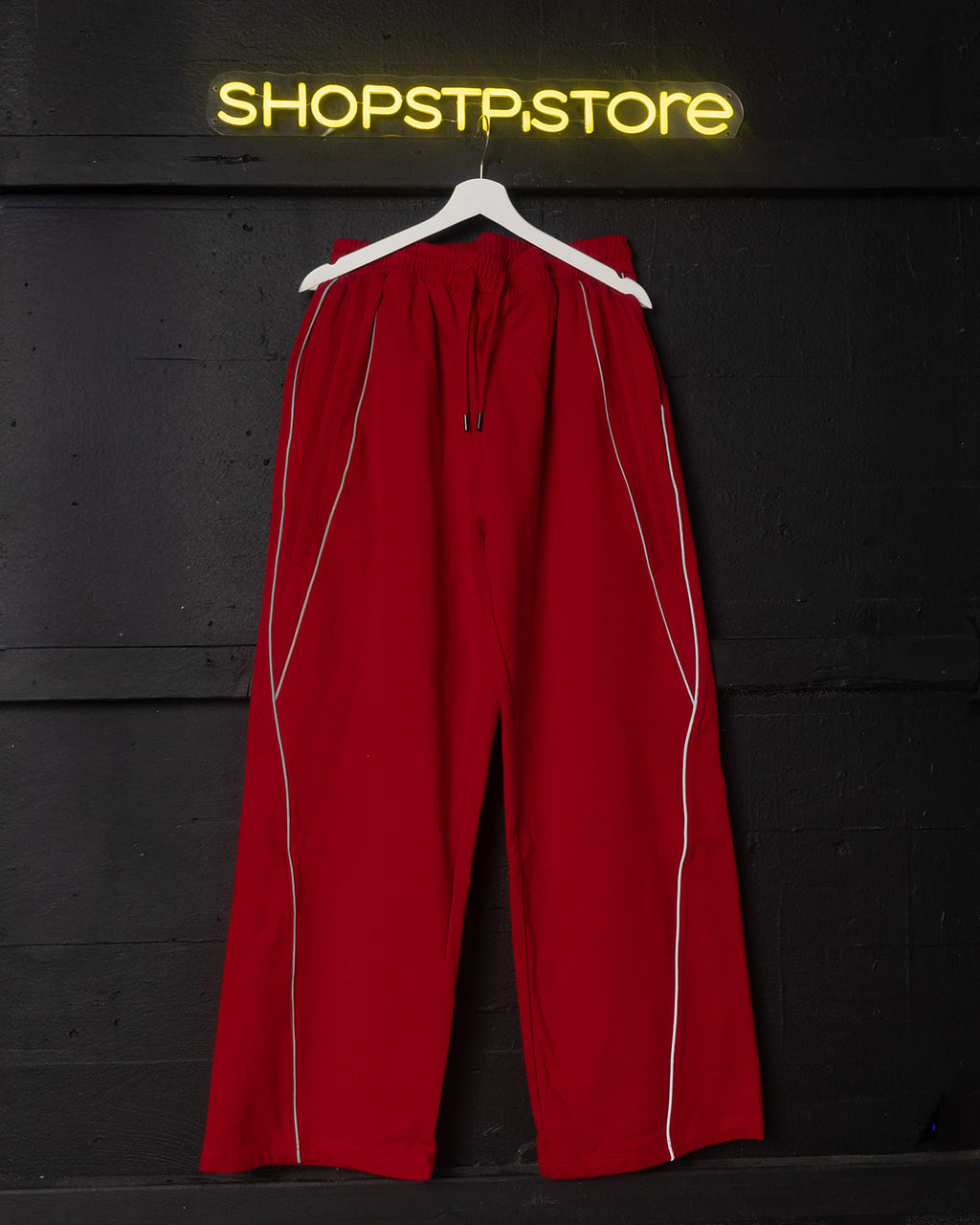 Red pants with white stripes on a hanger against a black background with 'SHOPSTP STORE' neon sign.
