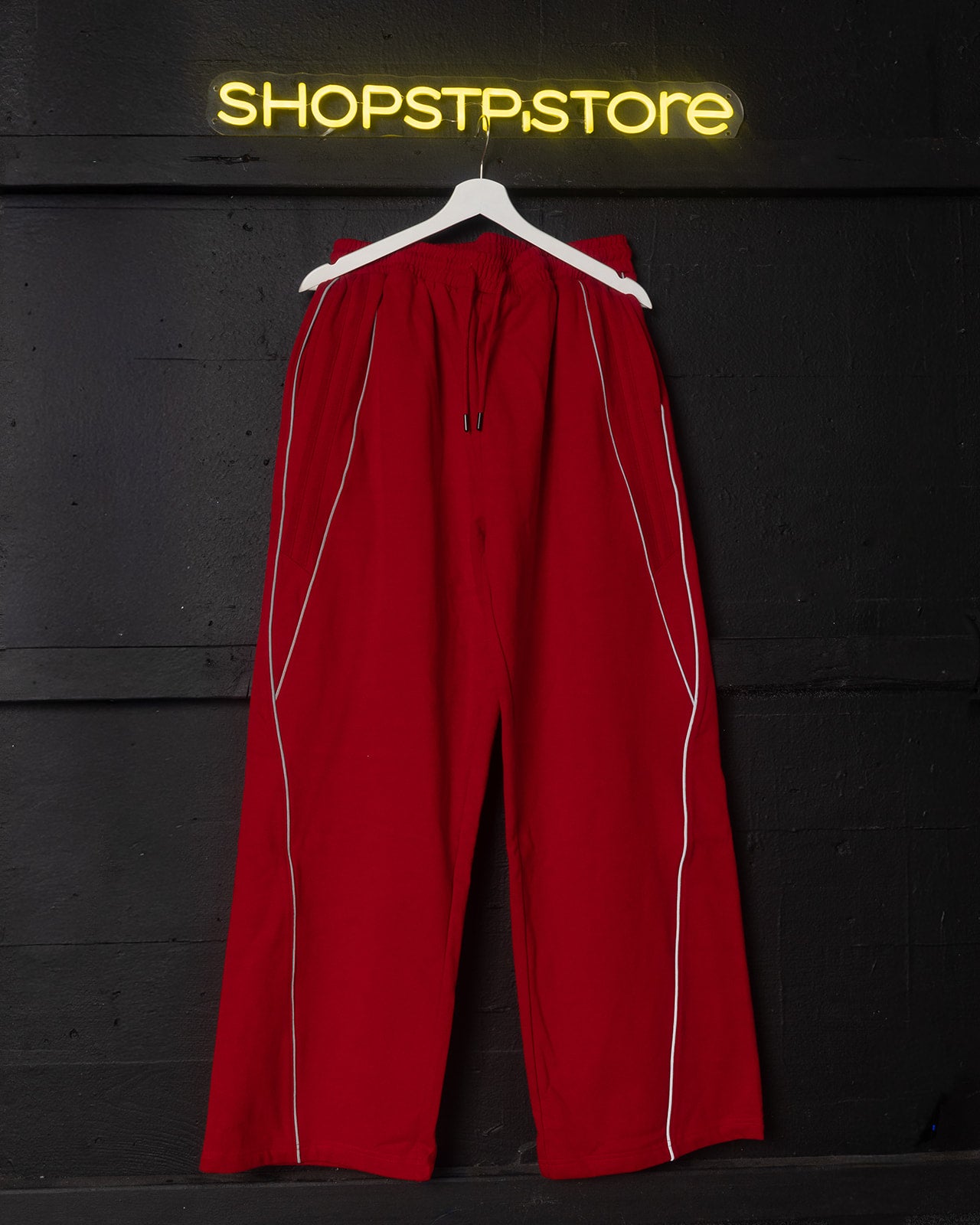 Red pants with white stripes on a hanger against a black background with 'SHOPSTP STORE' neon sign.