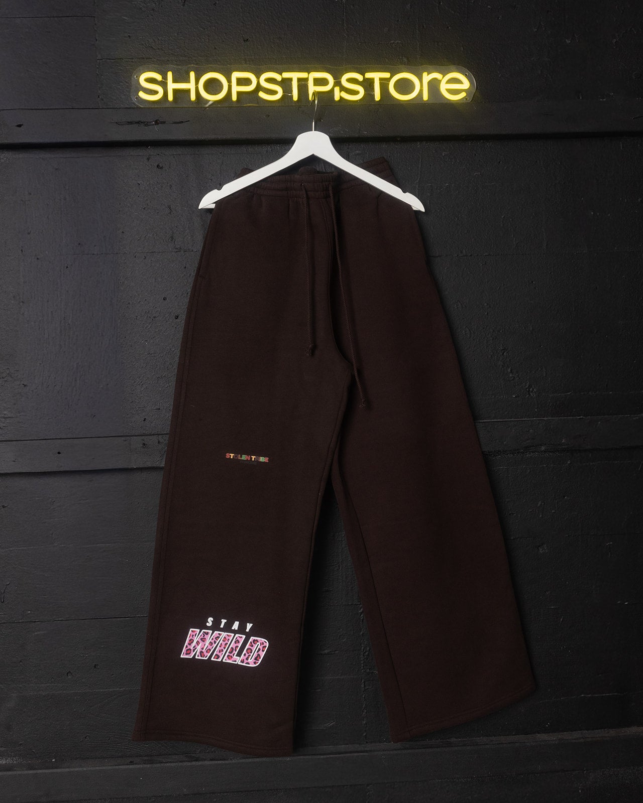 Brown pants hanging on a hanger with 'SHOPSTPSTORE' neon sign in the background
