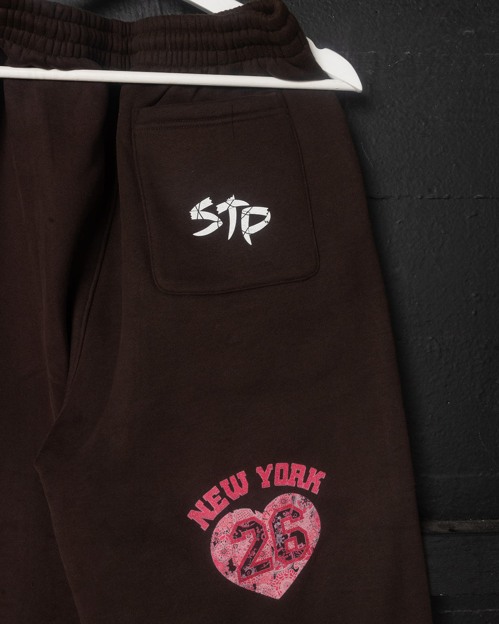 Brown sweatpants with a logo on the chest pocket and 'New York 26' design on the leg, against a dark background.