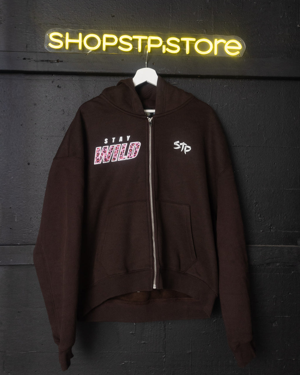 Brown hoodie with text design on a hanger against a dark wall with 'SHOPSTP.store' neon sign.