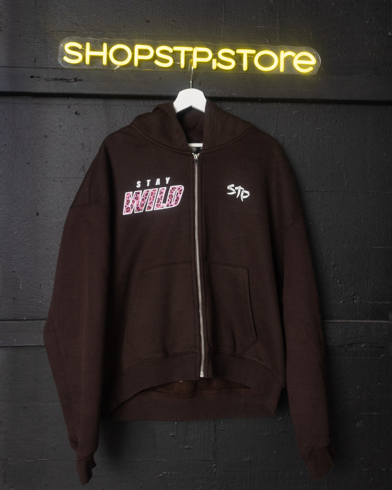 Brown hoodie with text design on a hanger against a dark wall with 'SHOPSTP.store' neon sign.