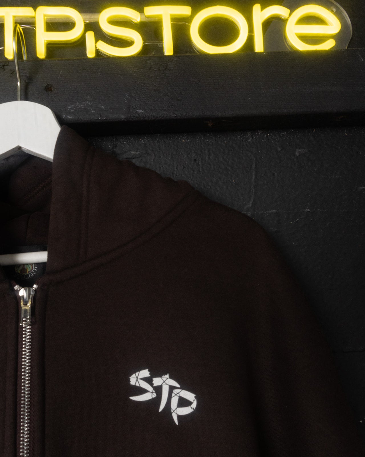 Brown hoodie with a logo in front of a neon sign reading 'STP Store' on a dark background