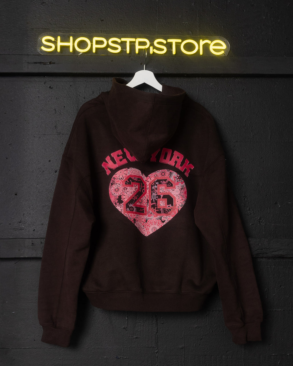 Brown hoodie with pink heart design and 'New York 26' text, hanging against a dark wall with 'ShopStpstore' neon sign.