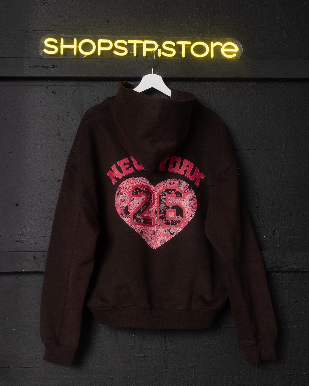Brown hoodie with pink heart design and 'New York 26' text, hanging against a dark wall with 'ShopStpstore' neon sign.