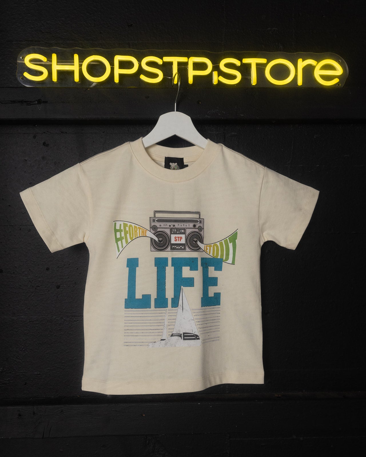 Beige t-shirt with graphic design and 'LIFE' text on a hanger against a dark background with 'SHOPSTP STORE' sign.