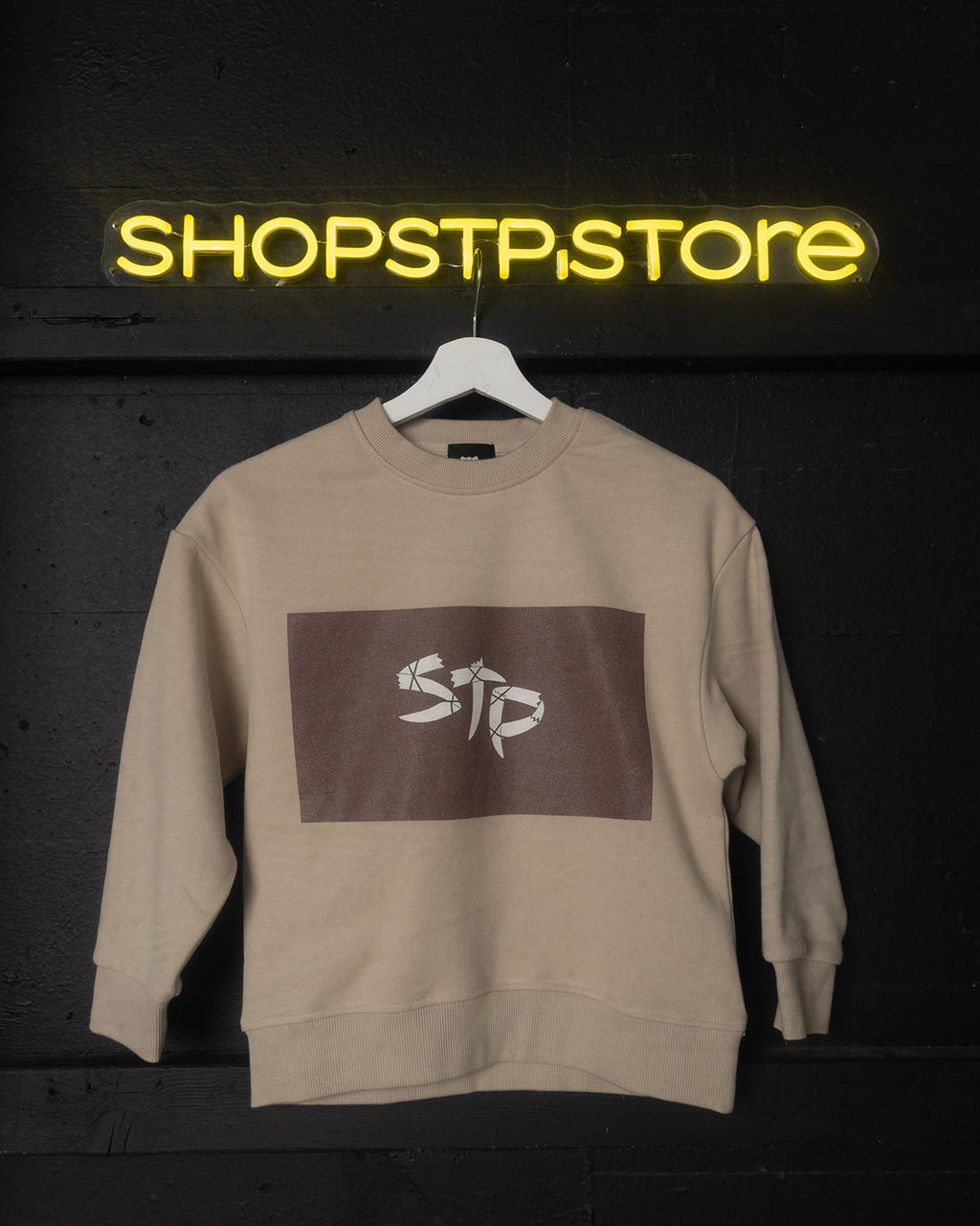 Beige sweatshirt with logo on a hanger against a dark background with 'SHOPSTP STORE' neon sign.
