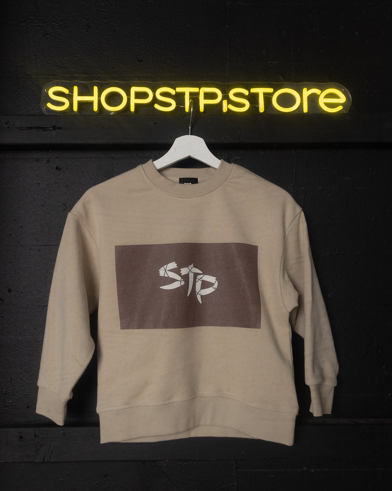 Beige sweatshirt with logo on a hanger against a dark background with 'SHOPSTP STORE' neon sign.