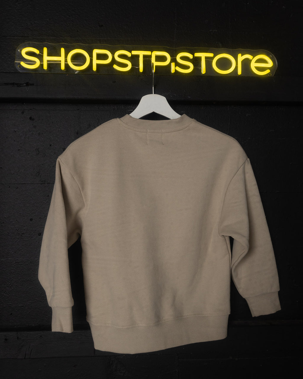 Beige sweatshirt on a hanger with 'SHOPSTPSTORE' neon sign in the background