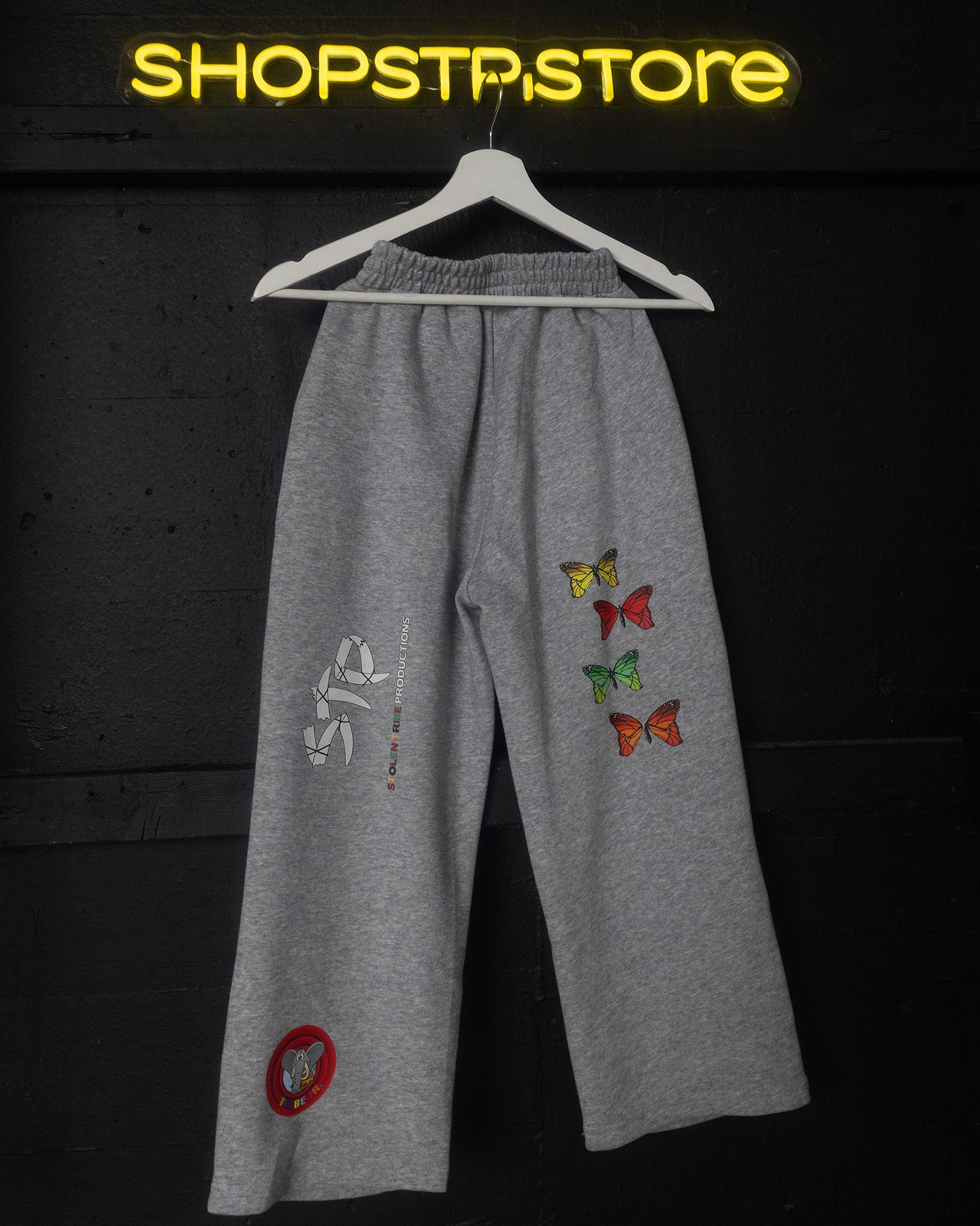 Gray sweatpants with colorful butterfly designs on a hanger against a dark background with 'SHOPSTP.STORE' branding.