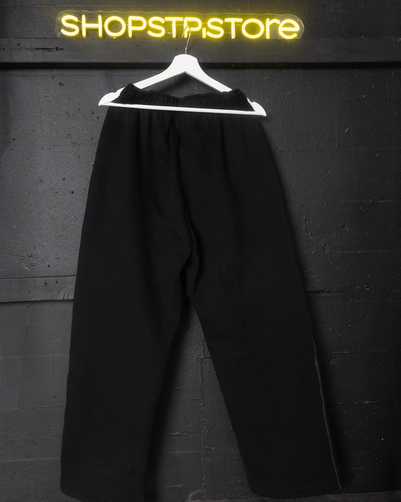 Black sweatpants on a hanger against a dark wall with 'ShopSTPstore' neon sign.