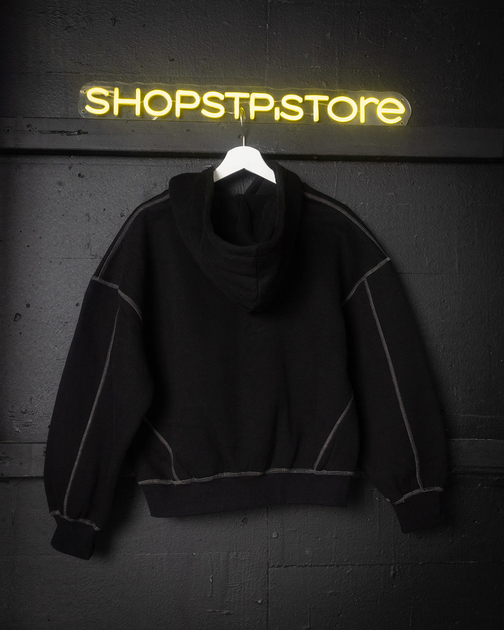 Black hoodie on a hanger with 'SHOPSTP.STORE' neon sign in the background
