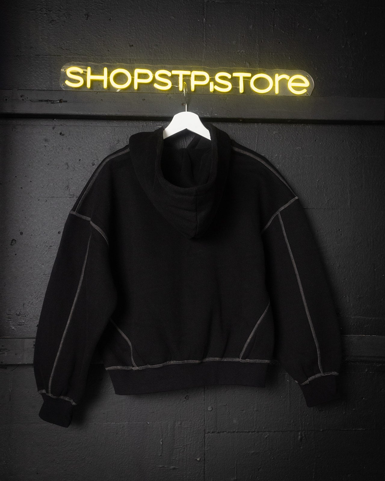 Black hoodie on a hanger with 'SHOPSTP.STORE' neon sign in the background