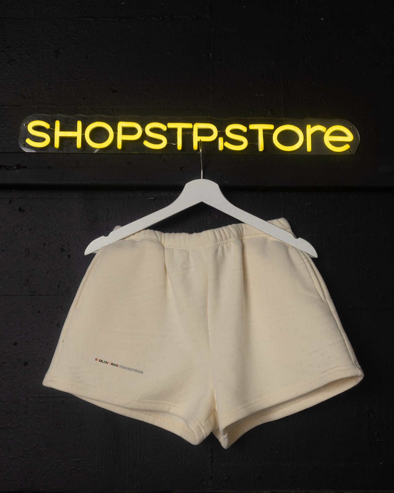 Beige shorts on a hanger with 'SHOPSTP.STORE' sign in yellow above on a black background
