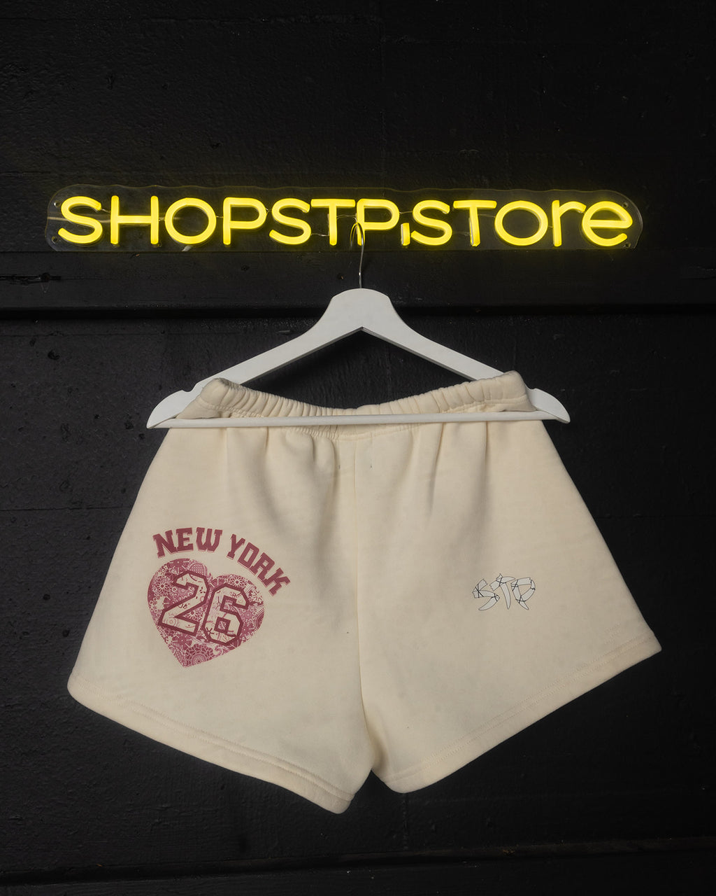 Beige shorts with 'New York 26' print on a hanger against a dark background with 'SHOPSTPSTORE' neon sign.