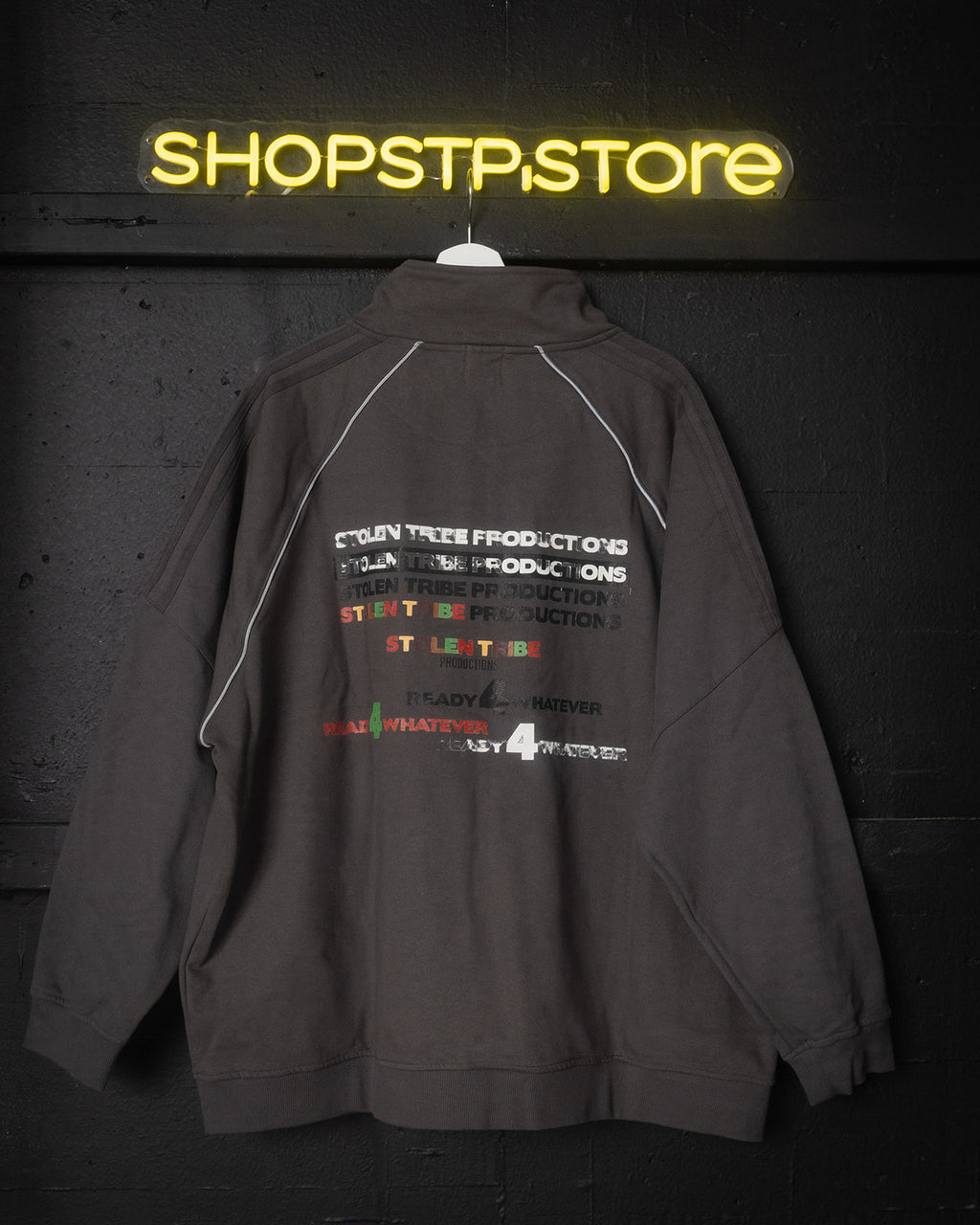 Gray jacket with text and logos on a dark background with 'SHOPSTPSTORE' neon sign.