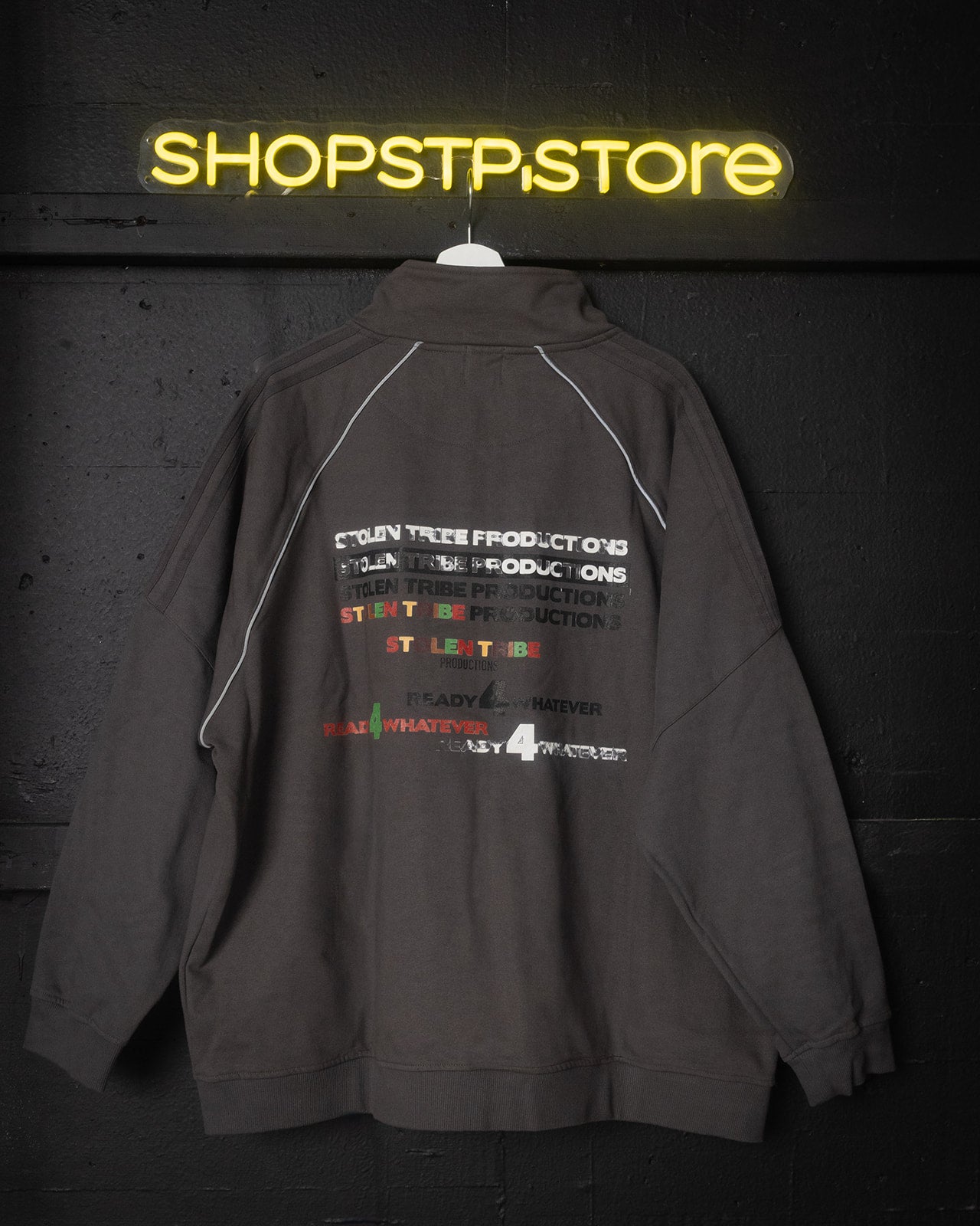 Gray jacket with text and logos on a dark background with 'SHOPSTPSTORE' neon sign.