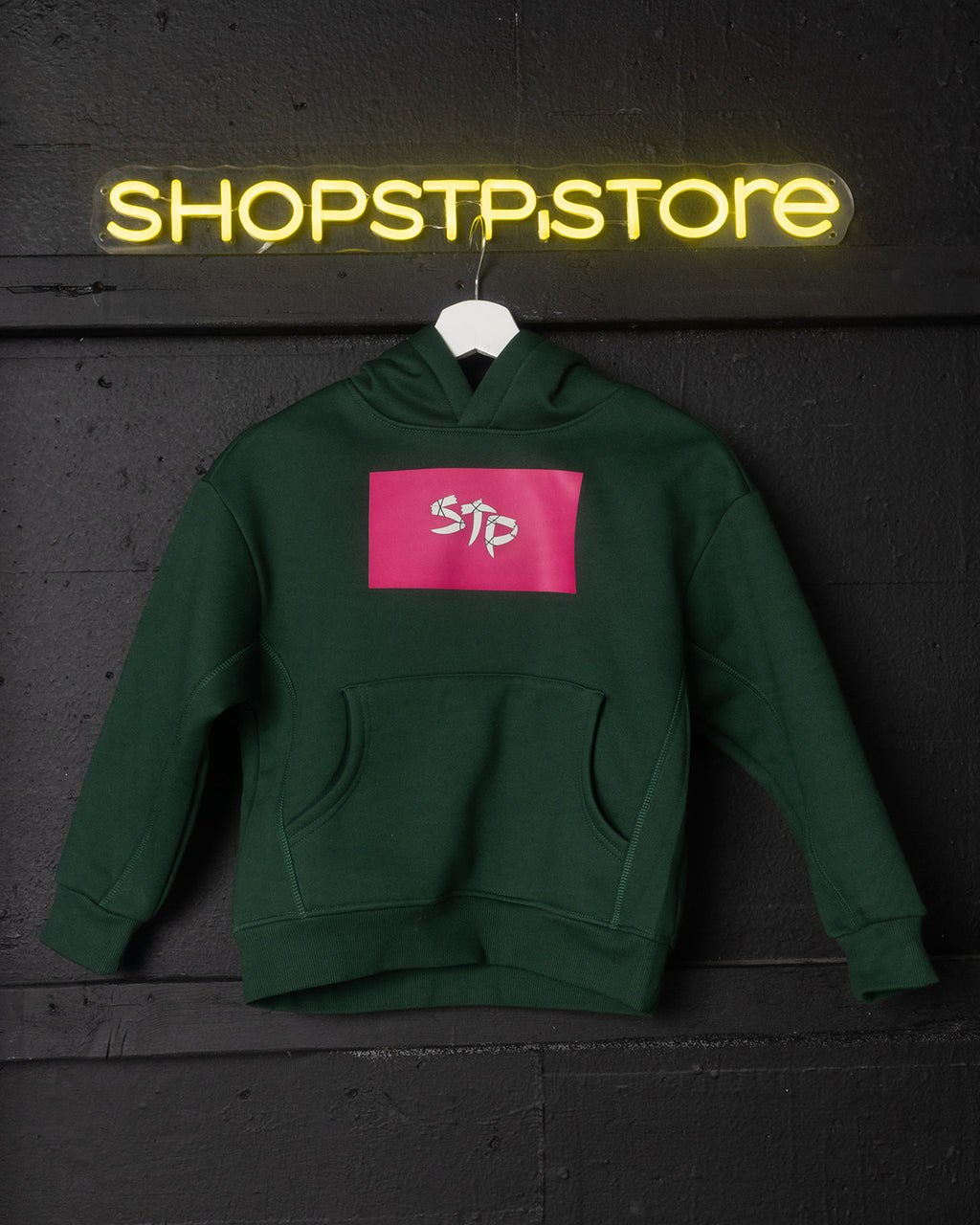 Green hoodie with pink logo on a hanger against a dark wall with 'SHOPSTP STORE' neon sign.