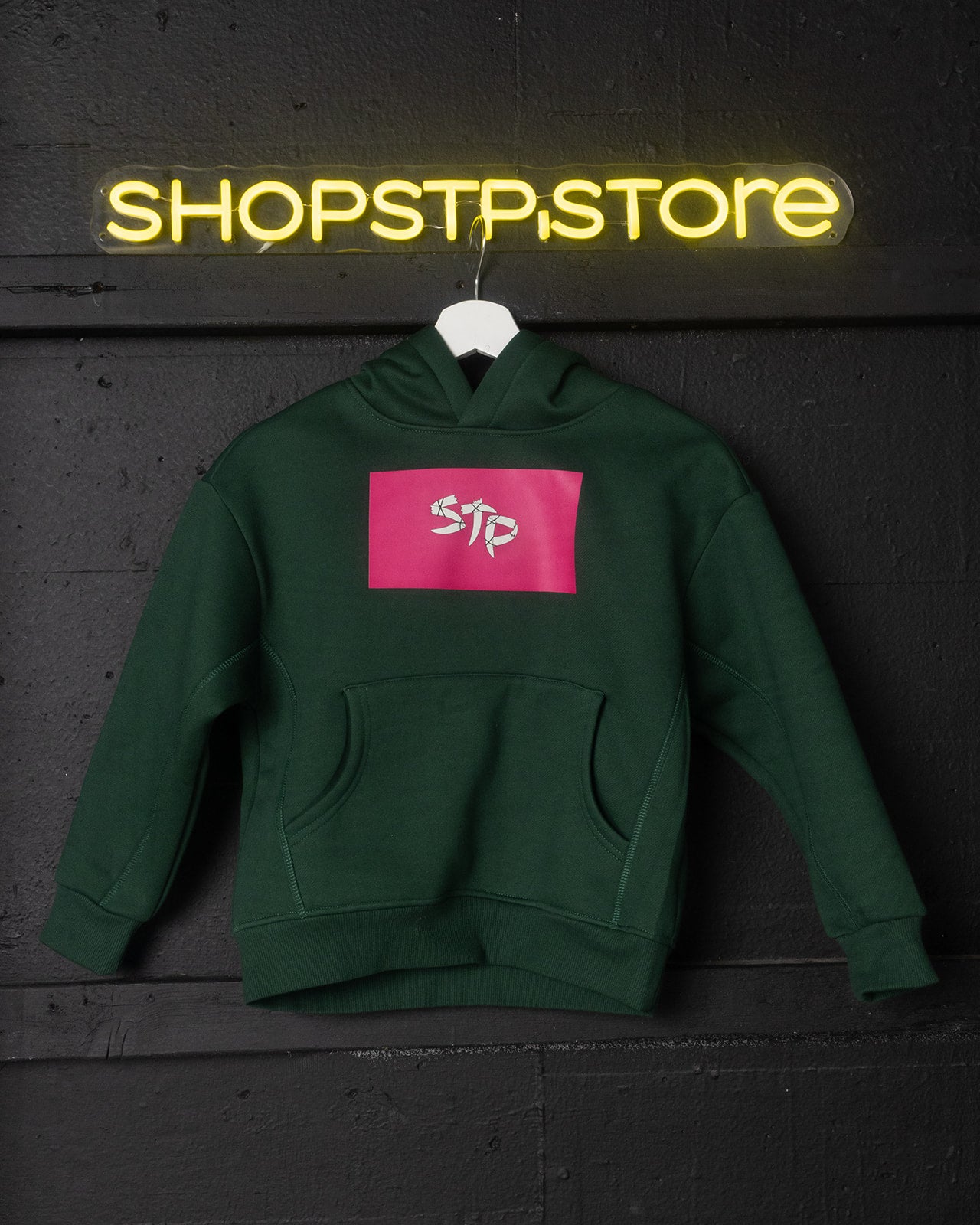 Green hoodie with pink logo on a hanger against a dark wall with 'SHOPSTP STORE' neon sign.