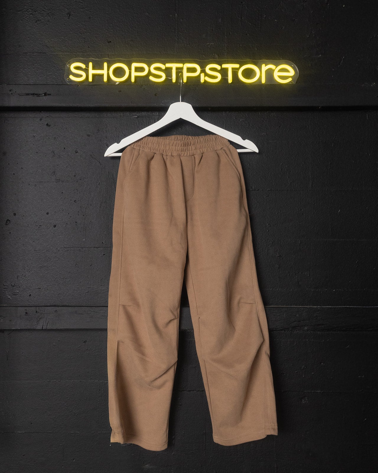 Brown pants on a hanger against a black wall with 'SHOPSTP STORE' neon sign.