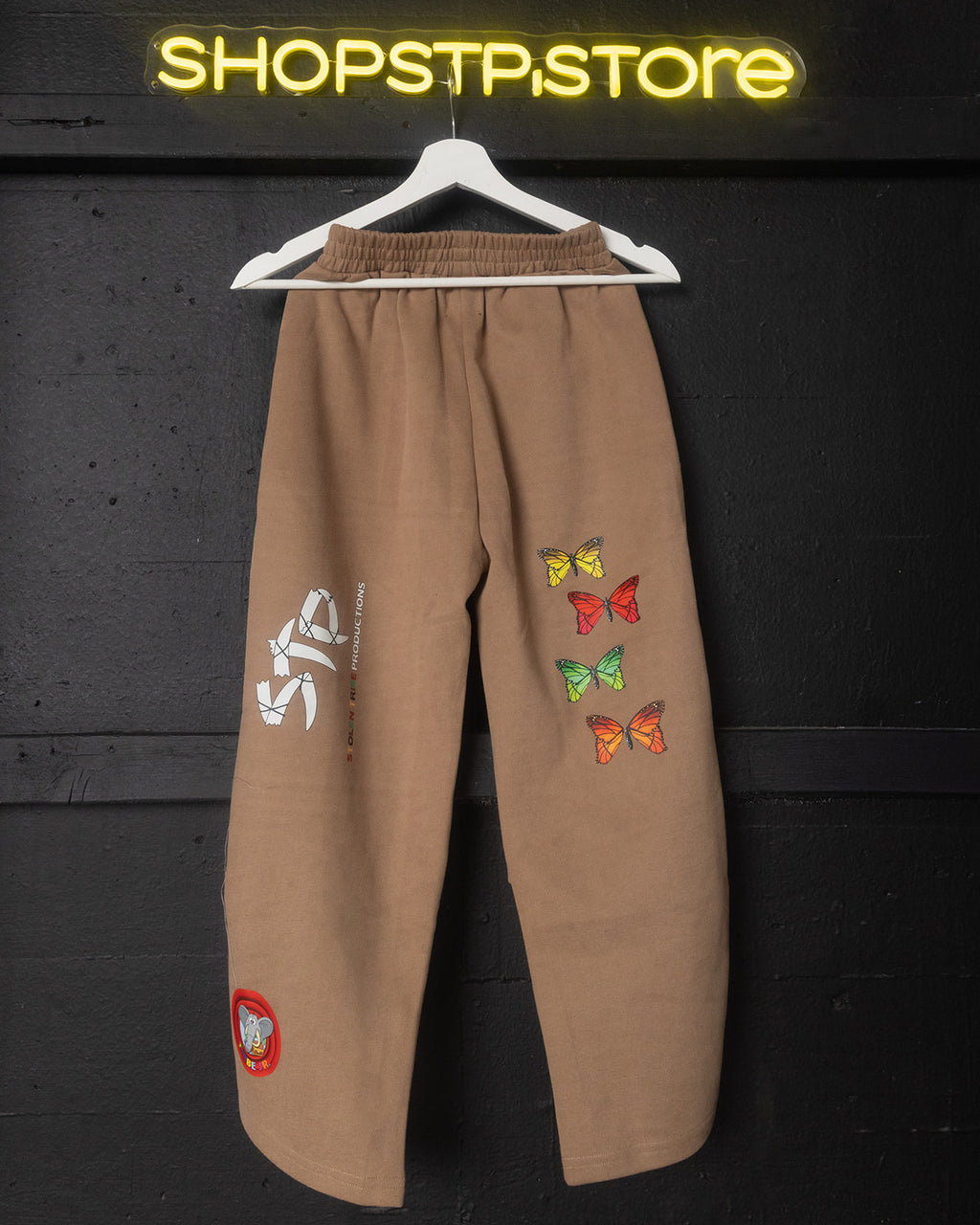 Brown pants with butterfly designs and patches on a hanger against a dark background with 'SHOPSTP.STORE' sign.