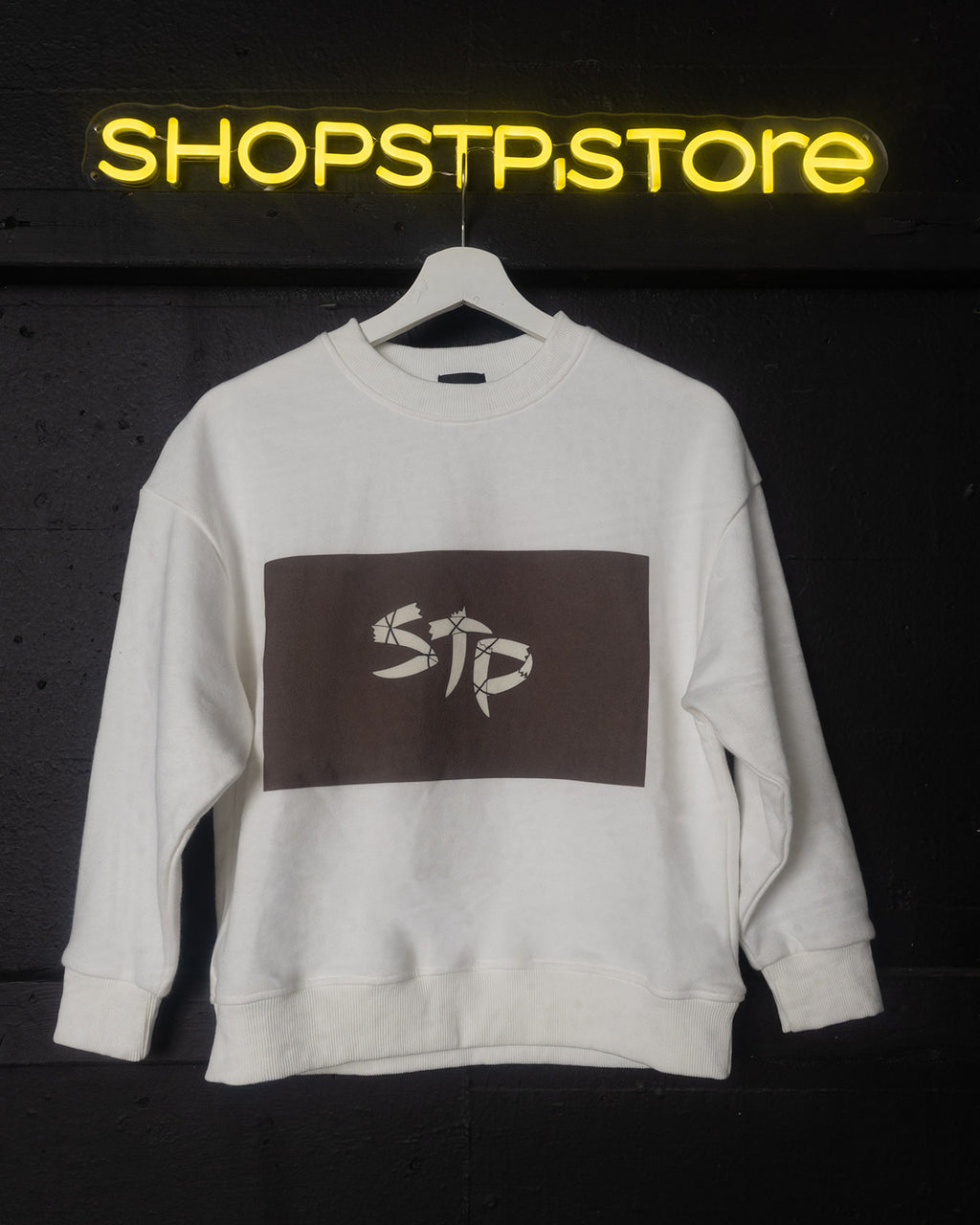 White sweatshirt with a logo on a hanger against a dark background with 'SHOPSTP STORE' neon sign.