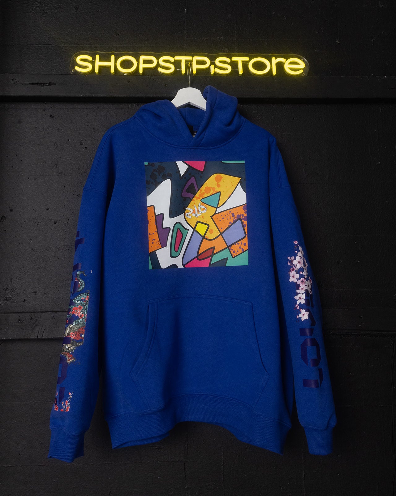 Blue hoodie with colorful abstract design on a hanger against a dark background with 'SHOPSTPSTORE' neon sign.