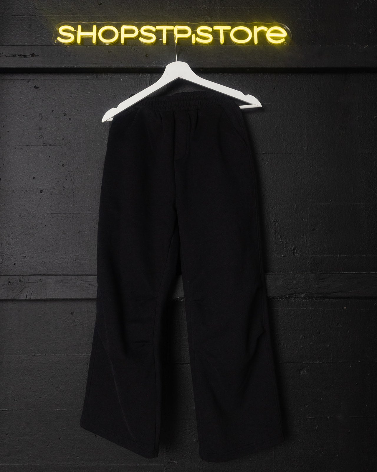Black pants on a hanger against a dark wall with 'SHOPSTPSTORE' neon sign.