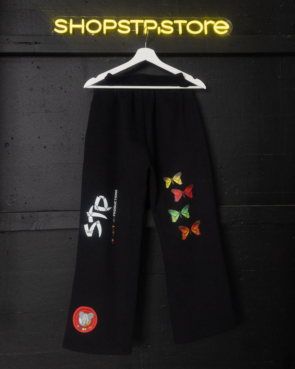 Black pants with colorful butterfly designs and a red patch on a hanger against a dark background with 'SHOPSTP STORE' neon sign.