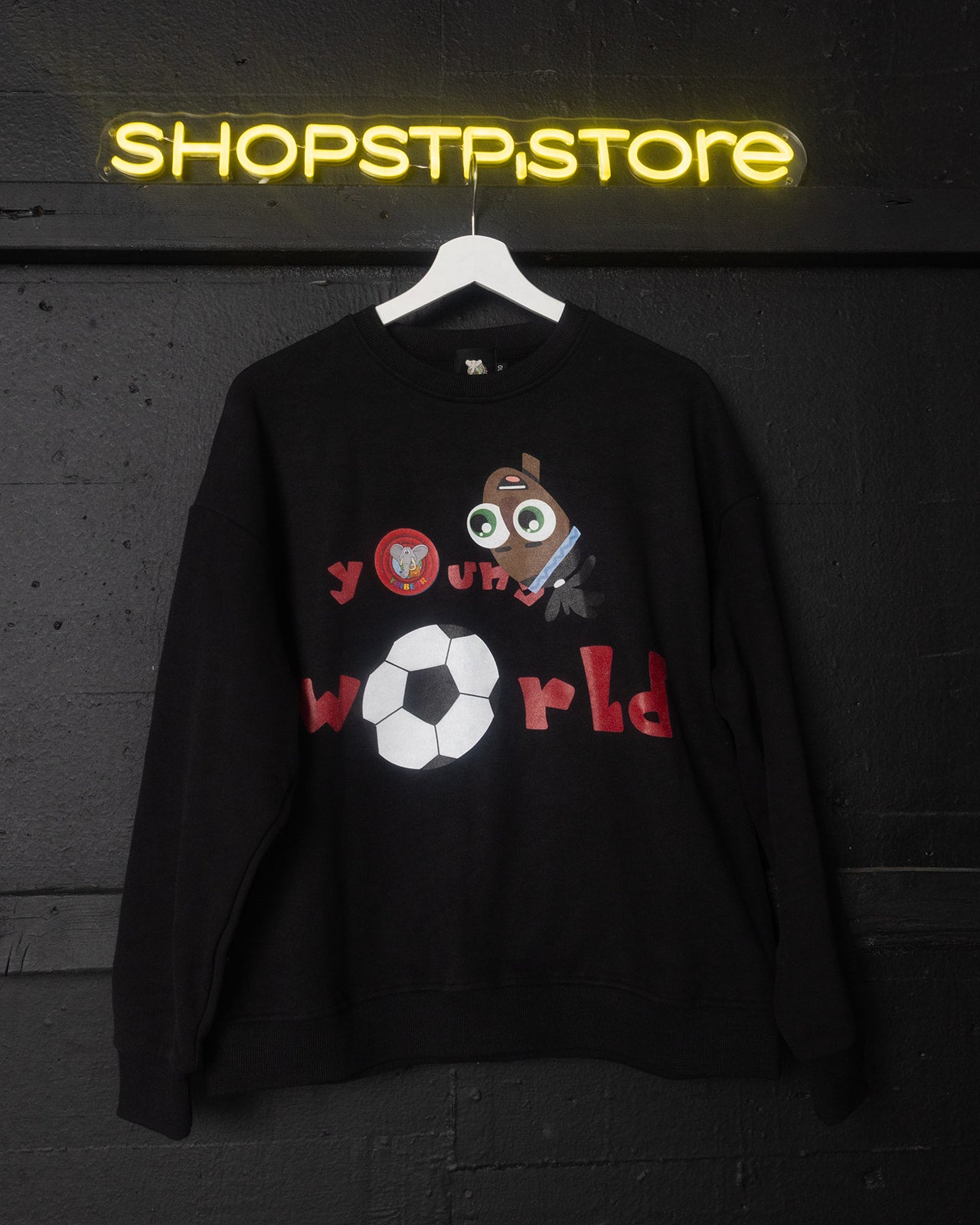 Black sweatshirt with cartoon character and text 'Young World' on a hanger against a dark background with 'SHOPSTPSTORE' neon sign.