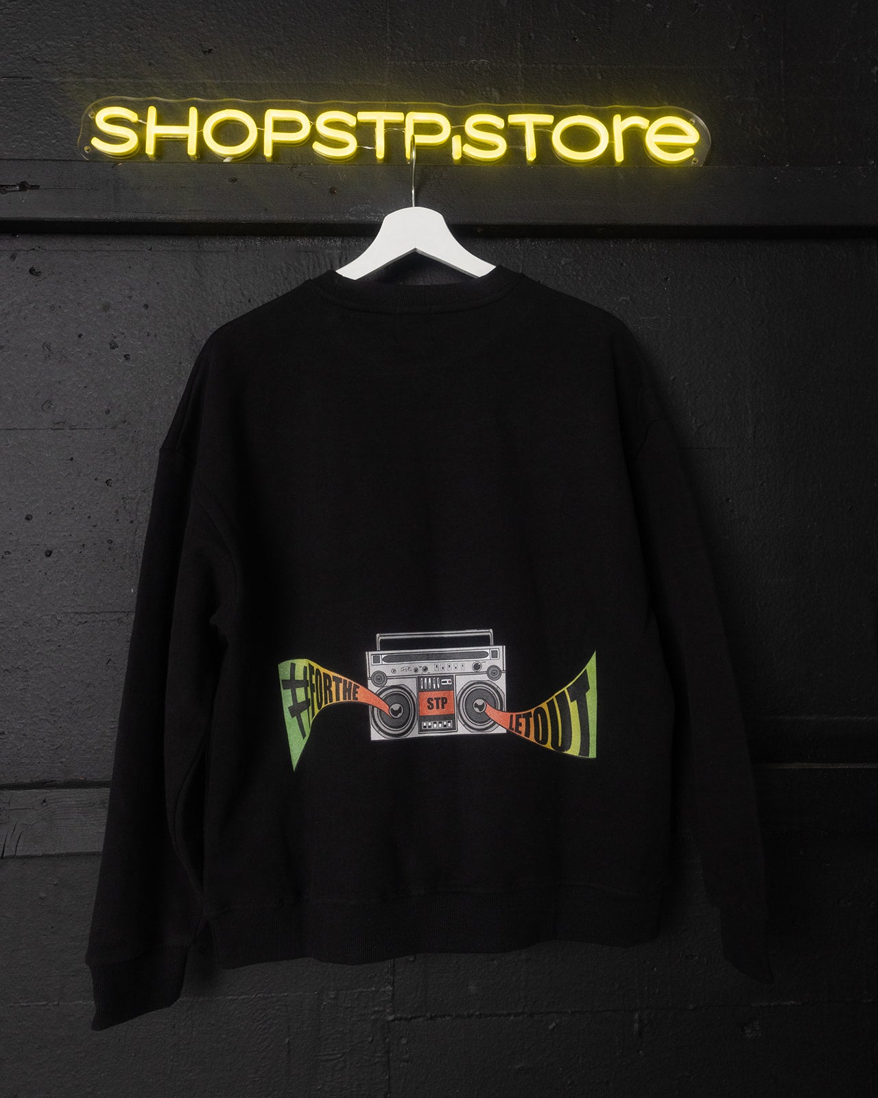 Black sweatshirt with a graphic design hanging in front of a neon 'SHOPSTPSTORE' sign.